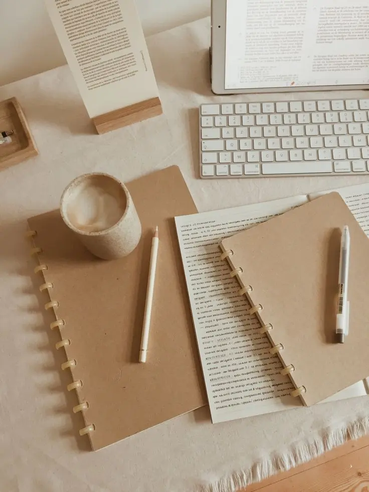 A cozy workspace setup with a tablet, wireless keyboard, two brown spiral notebooks, pens, and a cup of coffee on a beige tablecloth, creating a warm and minimal aesthetic.